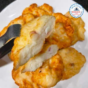 Cận cảnh đĩa chả mực chiên vàng ruộm, một miếng được cắt đôi cho thấy phần nhân mực tươi dai giòn bên trong, được giữ bằng kẹp kim loại trên đĩa trắng