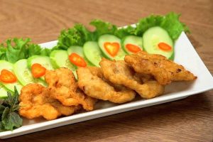 Đĩa chả mực giã tay chiên vàng ươm, bề mặt gồ ghề đặc trưng cho thấy rõ các hạt lựu mực dai giòn. Món chả mực truyền thống của Hải Sản Hoàng Ngư được bày trên đĩa trắng chữ nhật ăn kèm xà lách và dưa chuột.