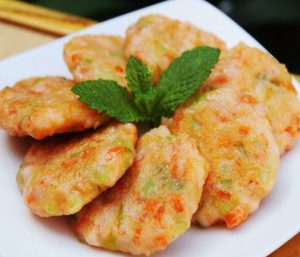 Cận cảnh đĩa chả mực tôm chiên vàng nhạt, nổi bật với những đốm thịt tôm hồng cam lấm tấm và điểm xuyết hành lá. Món chả tôm mực mềm xốp được bày trên đĩa sứ trắng trang trí lá bạc hà của Hải Sản Hoàng Ngư.